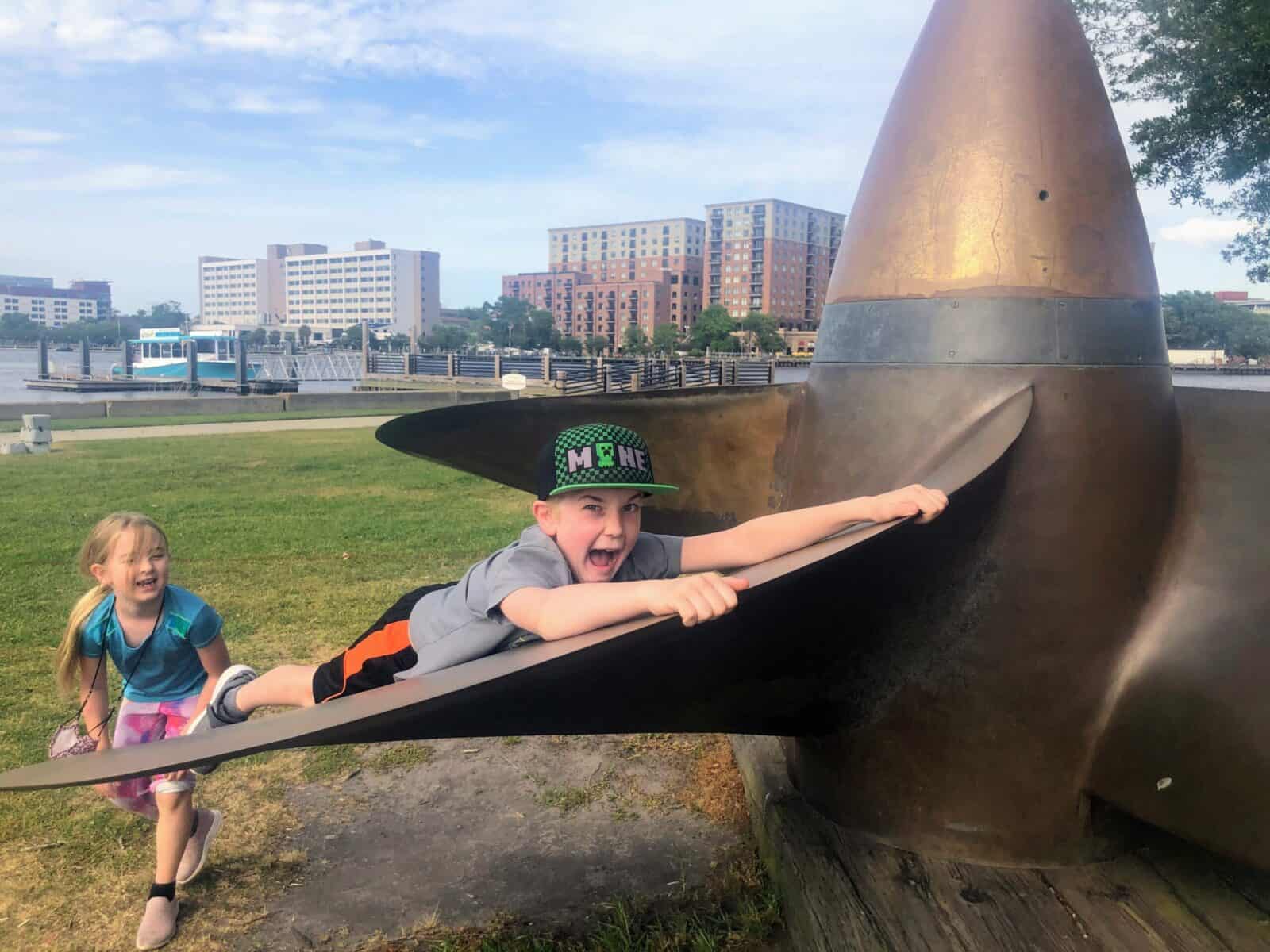 Children playing on a large outdoor sculpture in a park with city buildings in background.