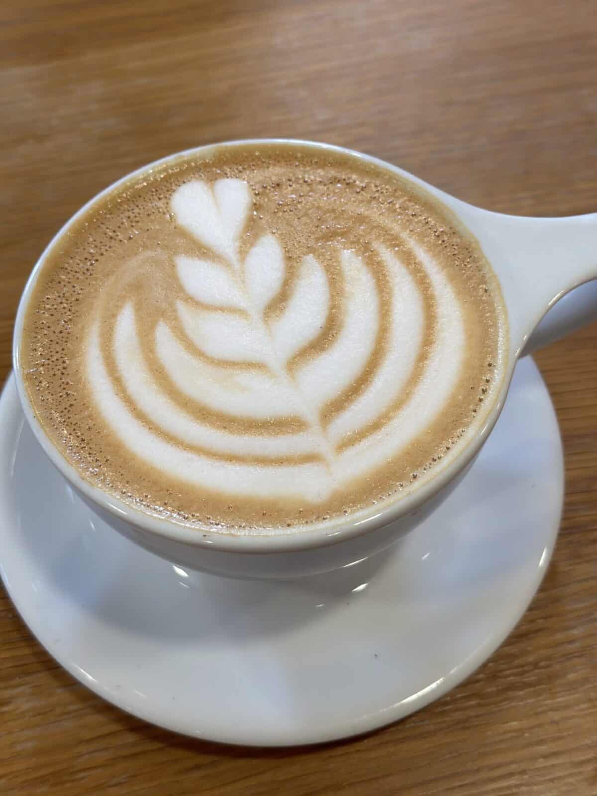A cappuccino with a leaf-pattern rosetta in the foam, served in a white cup and saucer on a light wood table at Casa Blanca Coffee Roasters.
