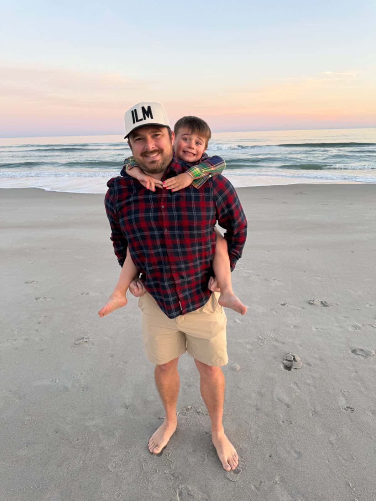 An adult in an ILM cap giving a child a piggyback ride on the beach at sunset near Wilmington, NC.