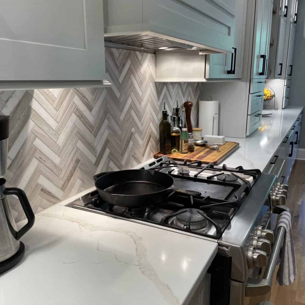 Marble herringbone backsplash behind a gas range with a cast iron skillet under a cabinet-style range hood