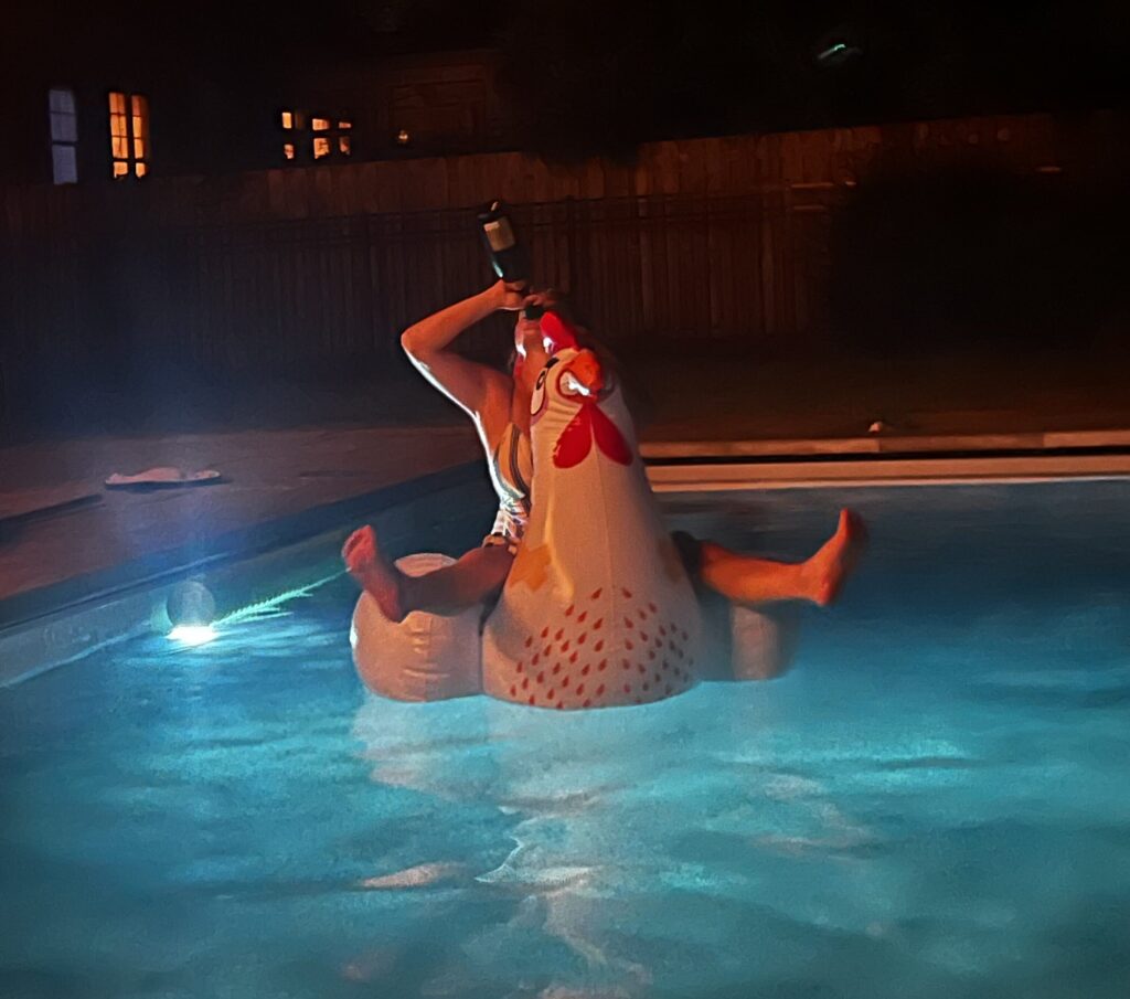 Floating on a giant chicken pool float at night with a glass of wine