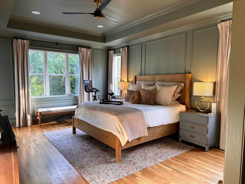 Primary bedroom after the makeover with green picture-frame molding walls, a warm wood bed, linen curtains and a bench under the window