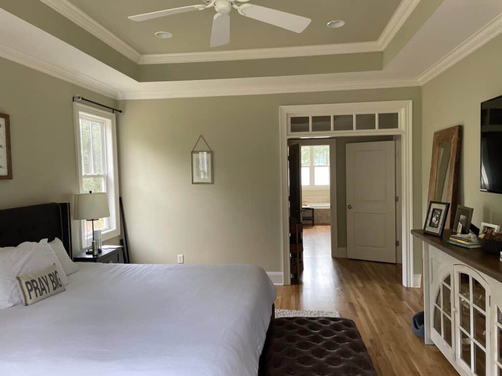 Primary bedroom before the makeover with a tray ceiling, plain sage-gray walls and a dark upholstered bed