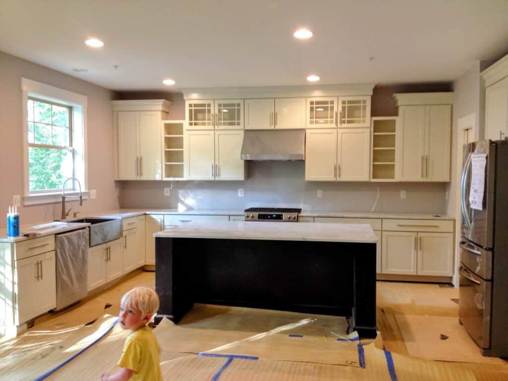 New-build kitchen near completion with cream shaker cabinets, glass-front uppers, a dark island, farmhouse sink and floors still covered in builder paper, with a toddler in a yellow shirt in the foreground